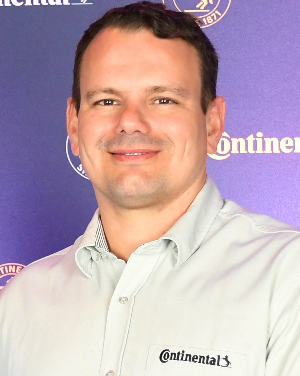 (foto perfil americano de Rodrigo Bonilha sorrindo com camisa cinza com bordado da Continental no bolso dianteiro no peito e fundo da foto com backdrop com logos da Continental - homem branco, sem barba, cabelo castanho penteado de lado)
