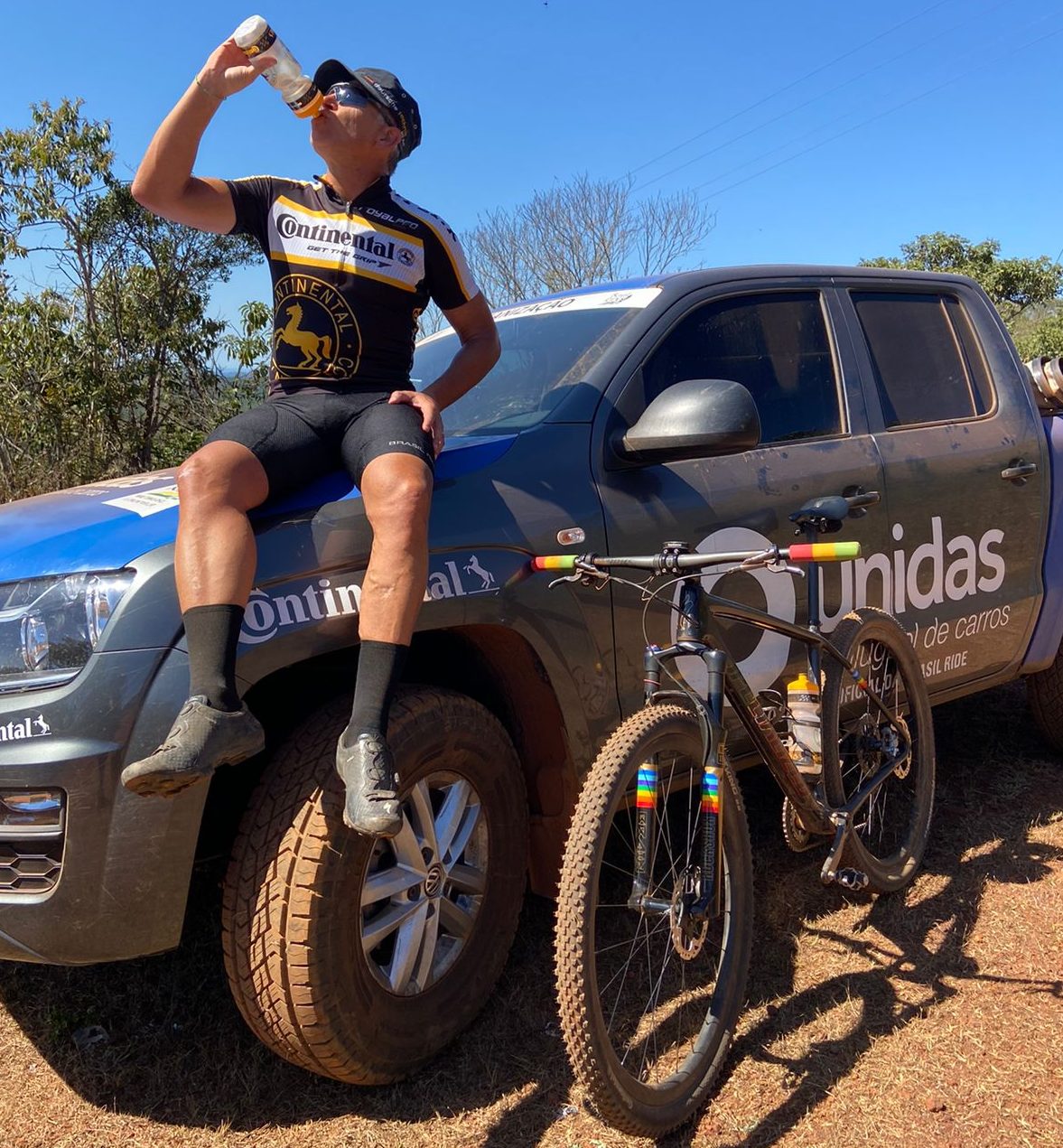 (foto de homem com rouba de ciclismo da Continental, boné e óculos, tomando agua em garrafa enquanto está sentado em caminhonete com pneu Continental e adesivada com Continental, enquanto bicicleta com pneu Continental está apoiada na lateral da caminhonete)