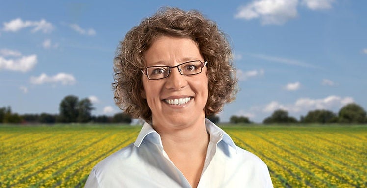 (Foto em perfil retrato de Carla Recker, com fundo de campo com plantação de dente-de-leão)