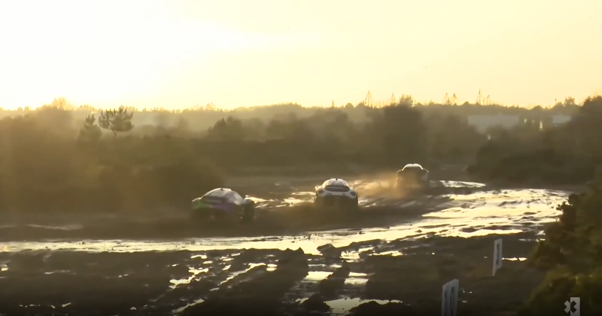 (imagem aberta de 3 veículos correndo em terreno com muita lama e por do sol durante corrida Extreme E em Dorset, Inglatterra)