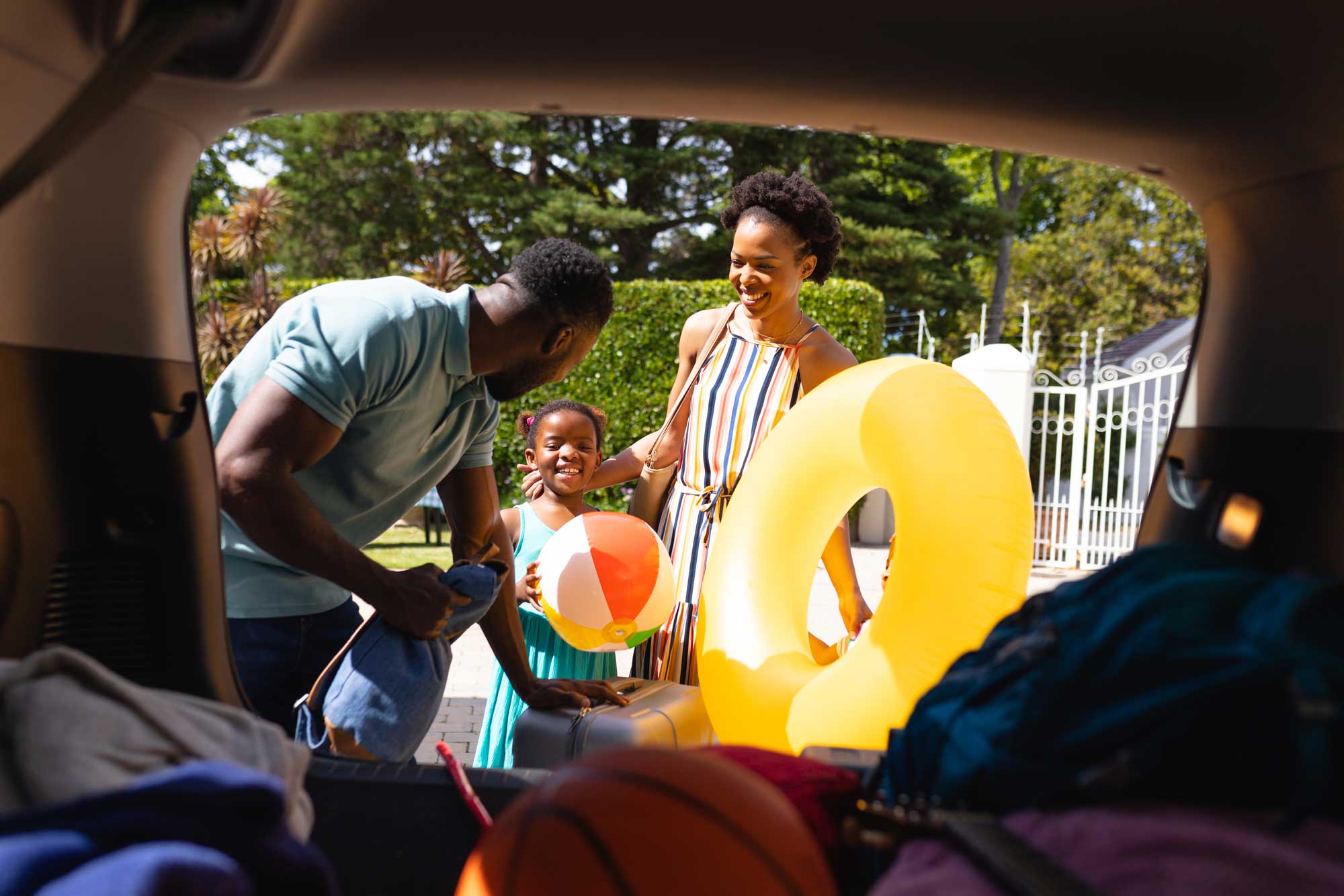 (imagem de família negra sorrindo enquanto guarda malas, bola e boia de praia em porta mala de SUV) 