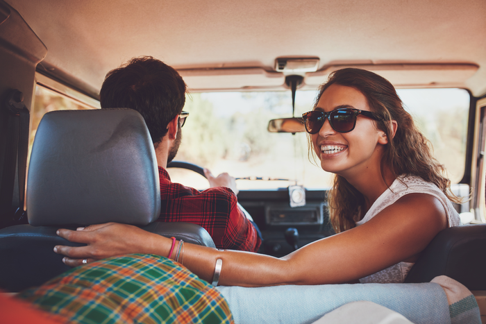 (imagem interna de veículo, com visão do banco traseiro, onde homem dirige o carro e mulher de óculos virada para trás sorri)