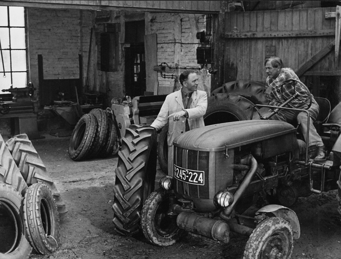 (imagem em preto e branco de dois homens ao lado de um trator antigo - oficina mecânica ao fundo, com pneus agrícolas ao lado)