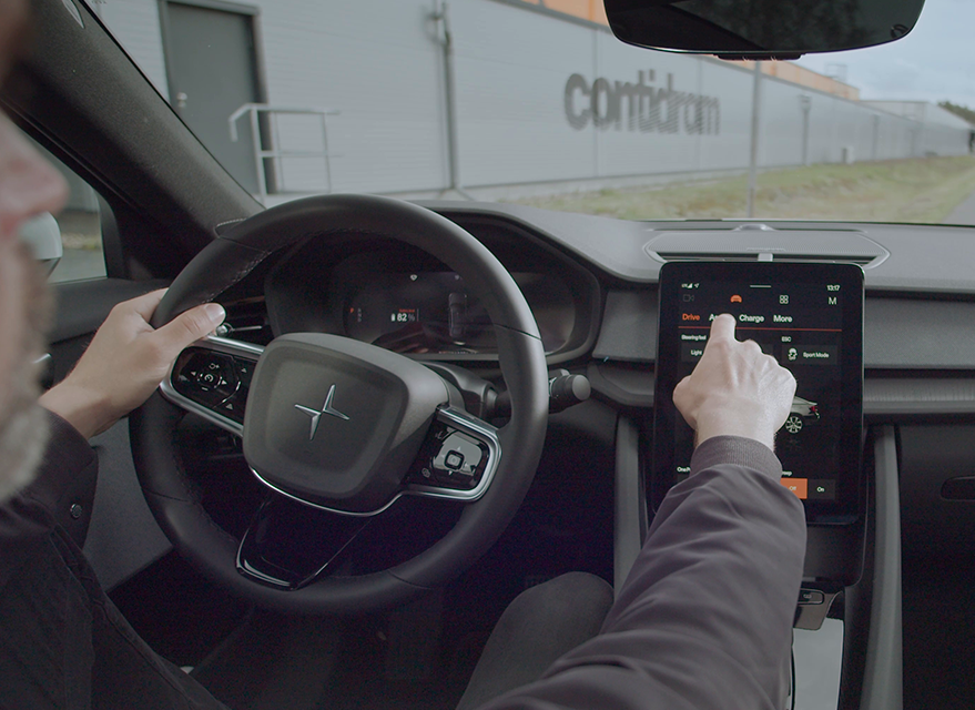 (foto interna de carro moderno andando na pista, mostrando pessoa com uma mão no volante e outra acessando painel digital do veículo)