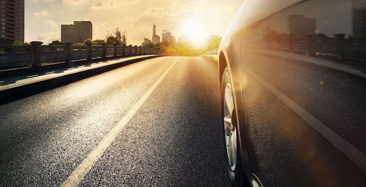 (imagem em close da lateral de carro com visão da traseira para a frente, em uma estrada asfaltada com prédios passando ao lado e por do sol com nuvens claras)