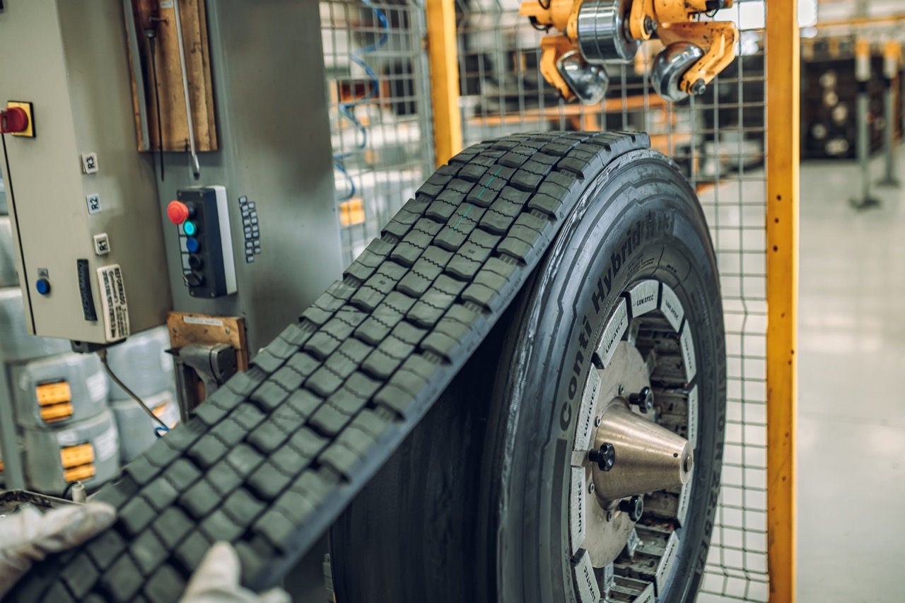 (imagem de mão com luva segurando banda de rodagem que está sendo aplicada em pneu de carga que se encontra em máquina)