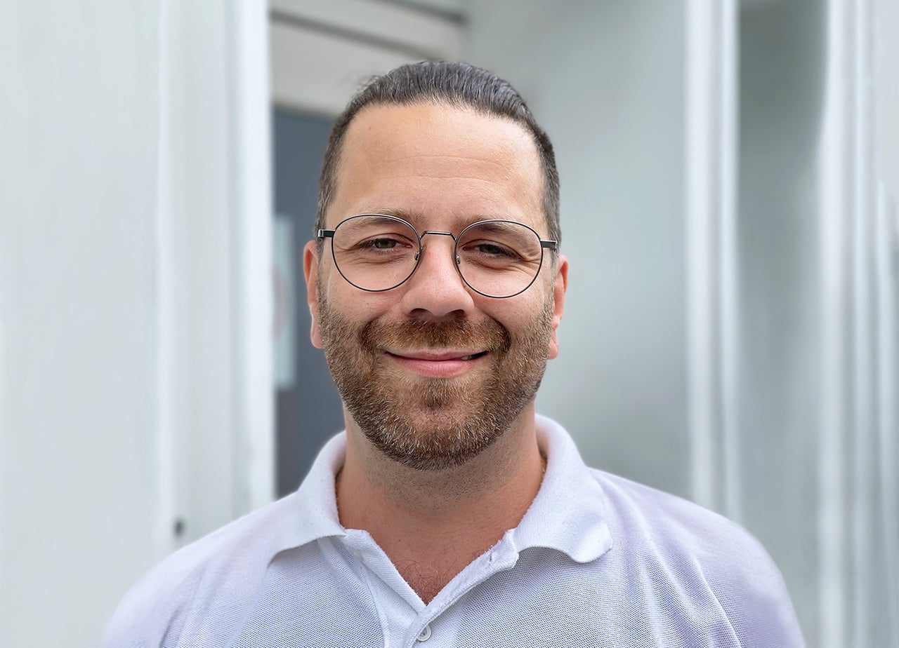 (imagem de homem branco sorrindo, com cabelos castanhos´penteados para trás, barba rala clara, olhos claros, óculos redondo e camiseta polo branca)