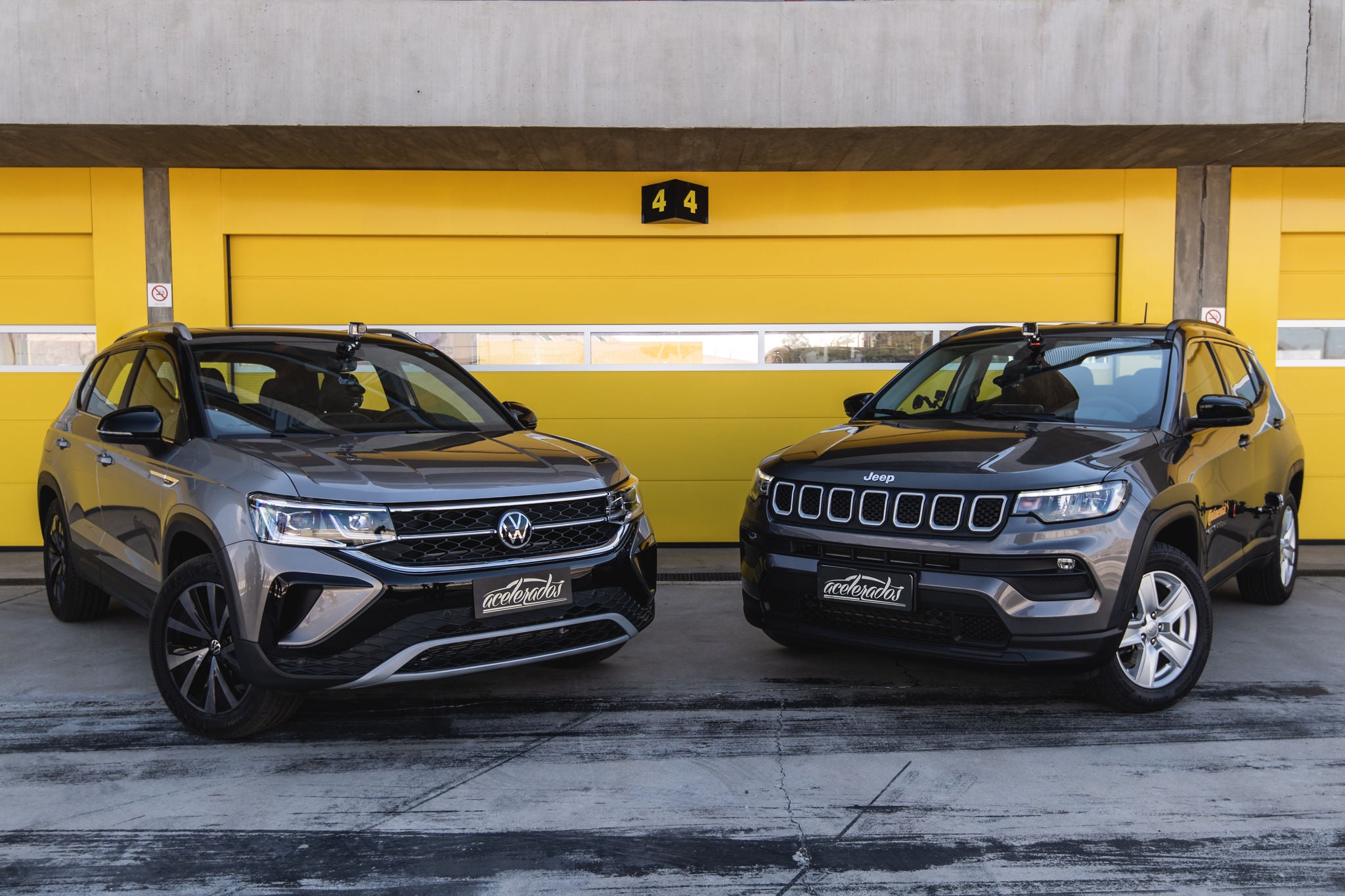 (foto frontal dos SUVs Taos da Volkswagen e Jeep Compass um ao lado do outro em frente a garagem amarela)