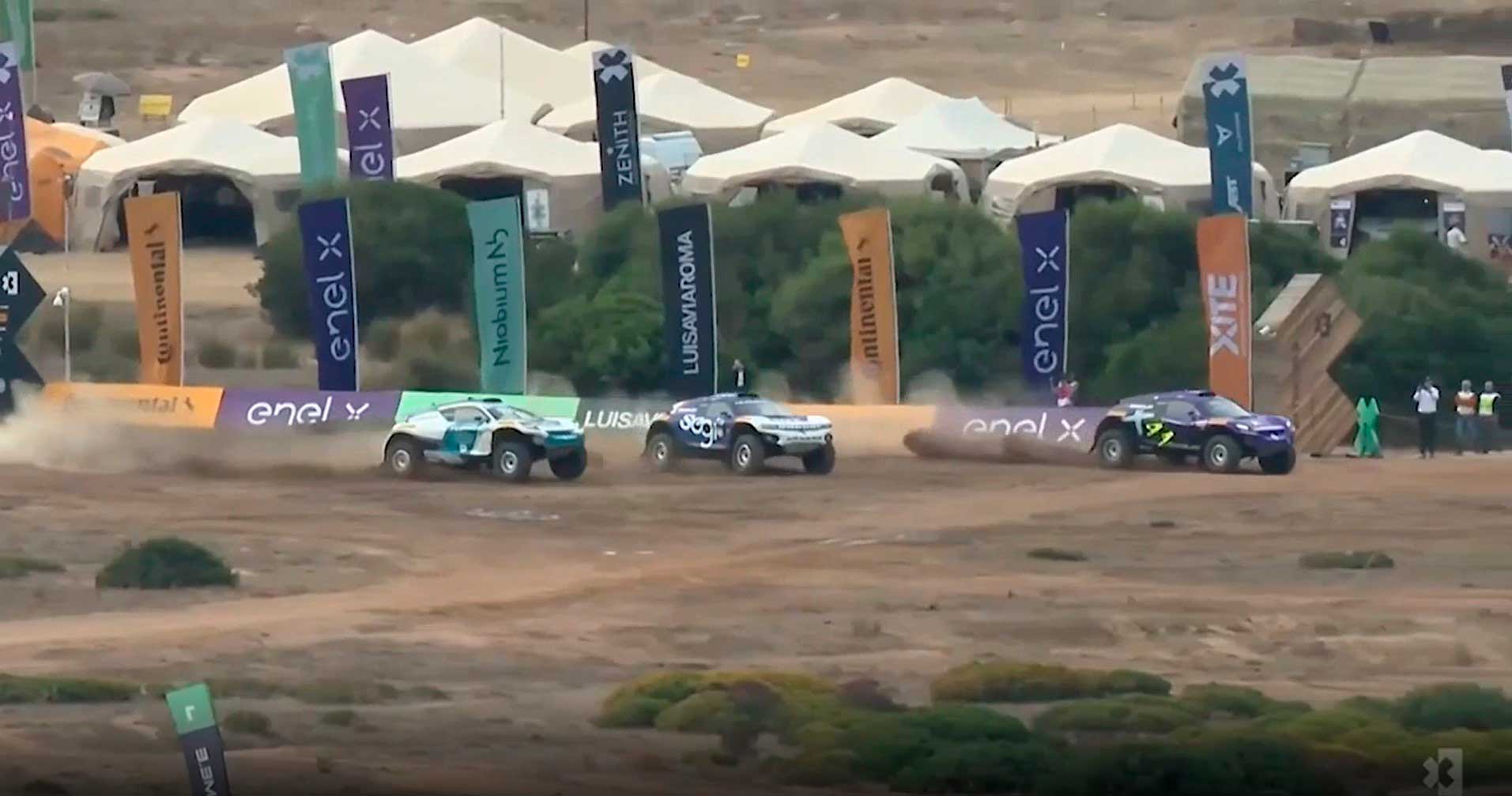 (imagem aberta de 3 veículos correndo em corrida do Extreme E em Sardinha Itália, com bandeiras e tendas no cenário de fundo)