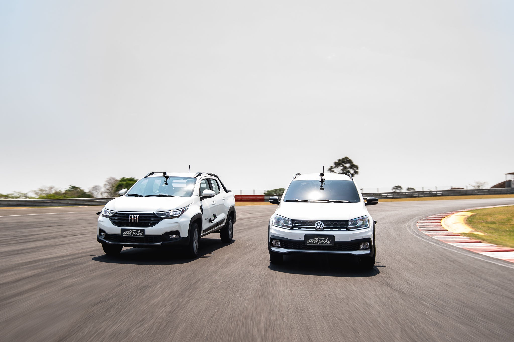 (foto de frente das picapes brancas da Volkswagen e Fiat, Saveiro e Nova Strada, calçadas com pneu Continental e andando na pista uma ao lado da outra)