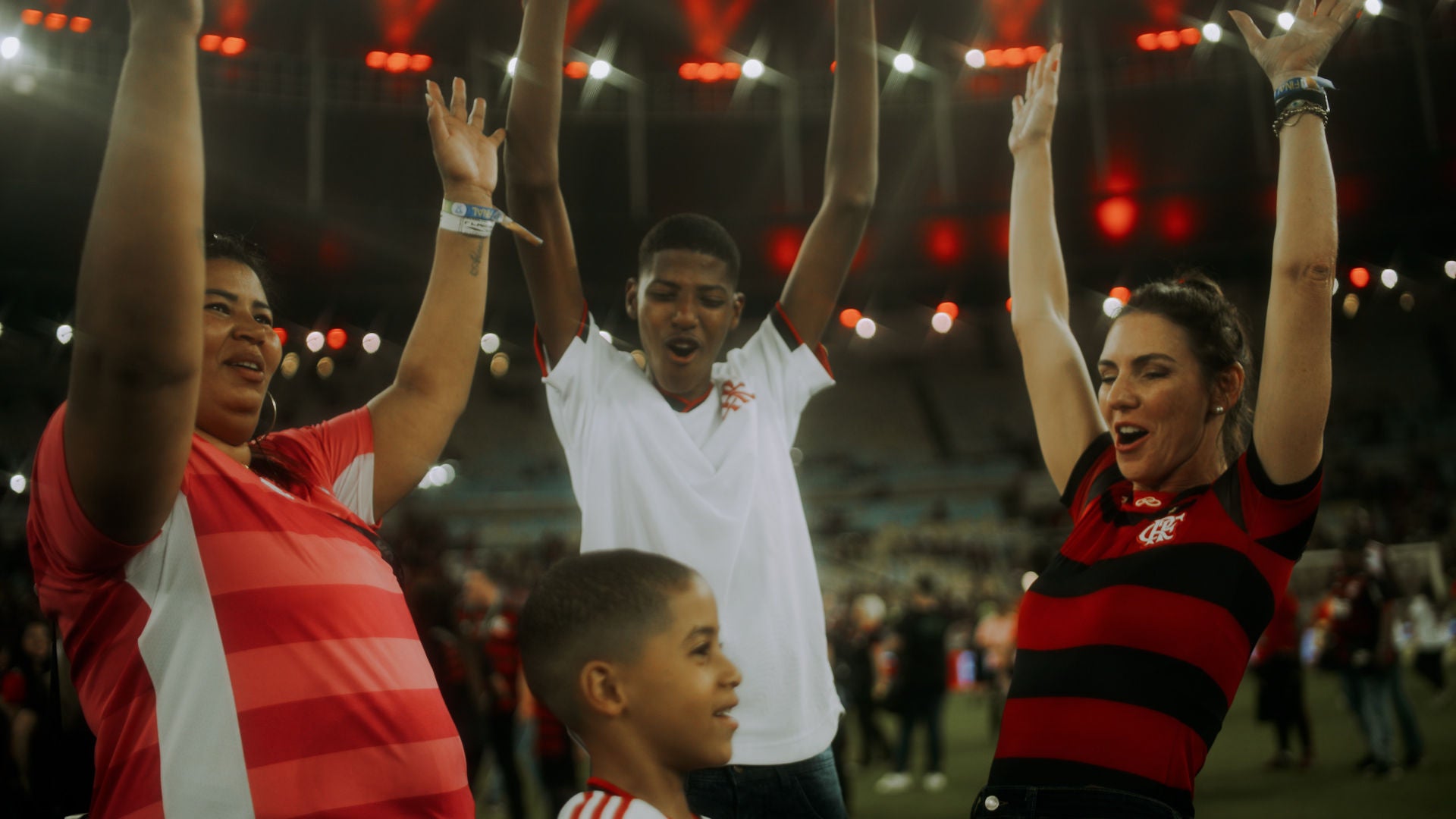 (foto de mãe Valéria, filho mais velho Alecsander e filho mais novo Arthur, vestidos com camisa do time Flamengo, torcendo com as mãos para cima junto a Glenda Kozlowski também vestida com a camisa do time)