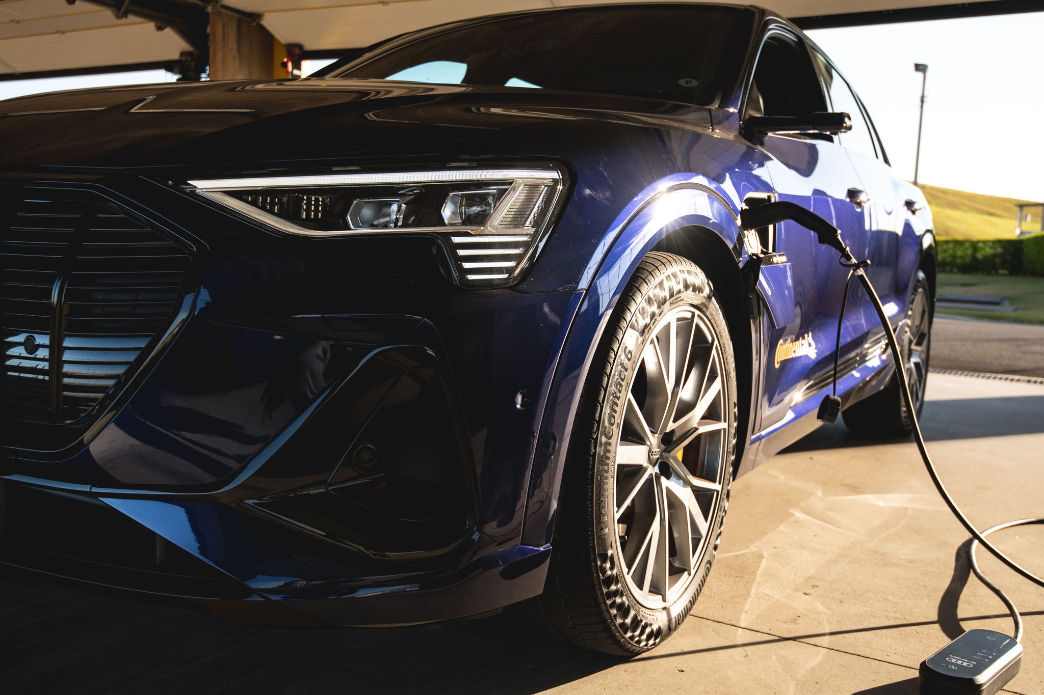 (foto frontal de SUV elétrico Etron azul da Audi, com pneus originais da Continental, sendo recarregado em tomada específica)