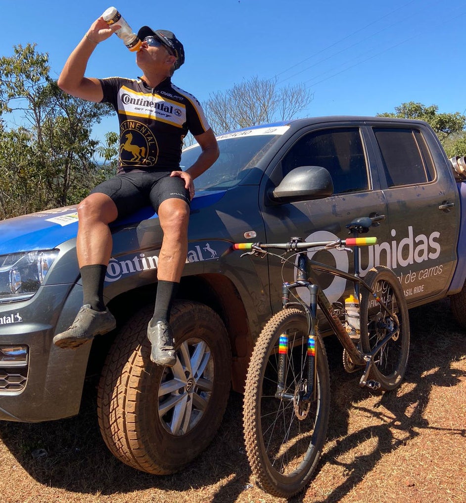 (foto de homem com rouba de ciclismo da Continental, boné e óculos, tomando agua em garrafa enquanto está sentado em caminhonete com pneu Continental e adesivada com Continental, enquanto bicicleta com pneu Continental está apoiada na lateral da caminhonete)