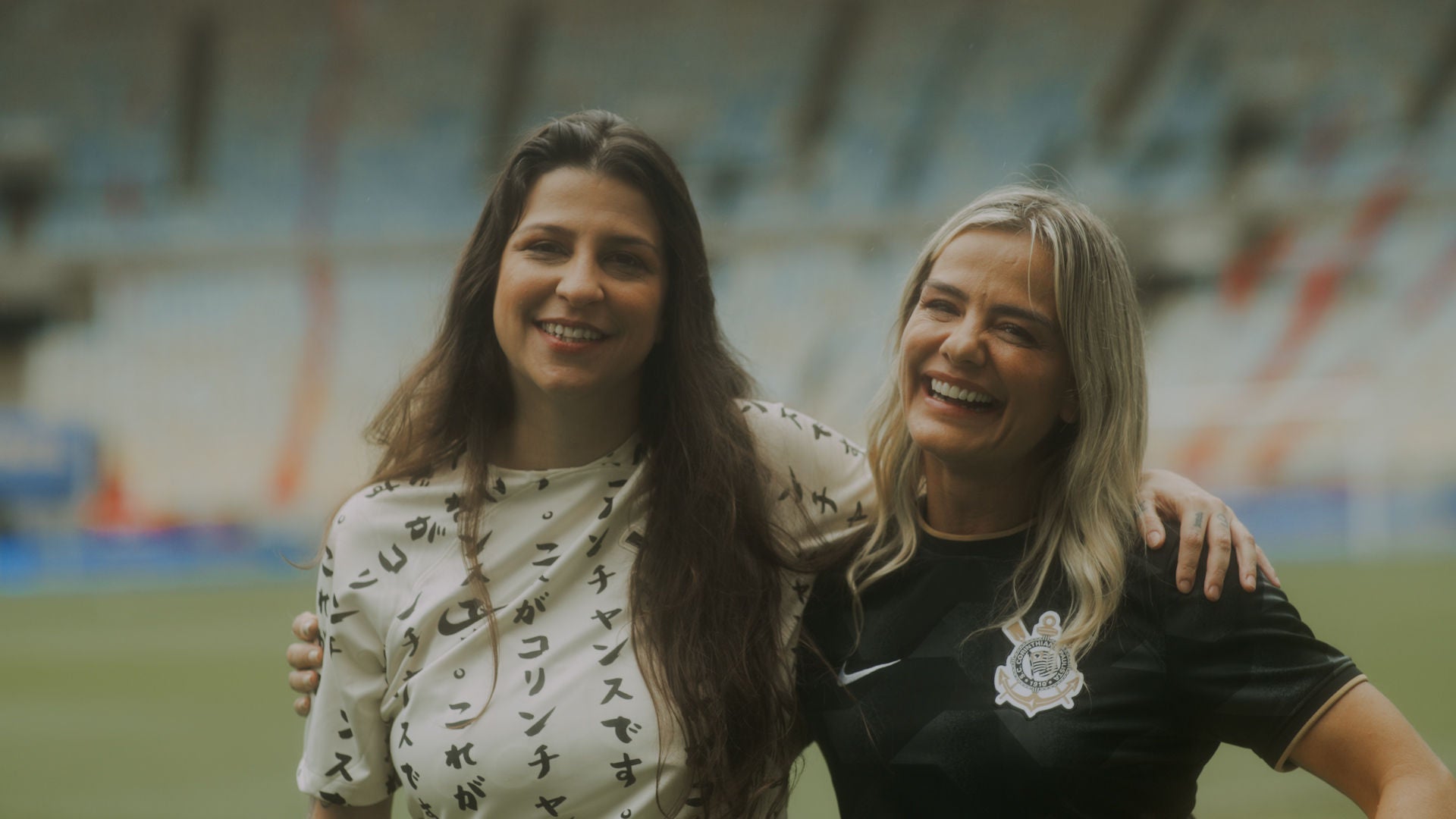 (foto de frente da corinthiana Leonor Macedo com camisa do Corinthians, abraçada e sorrindo junto da comentarista esportiva Milena Domingues)