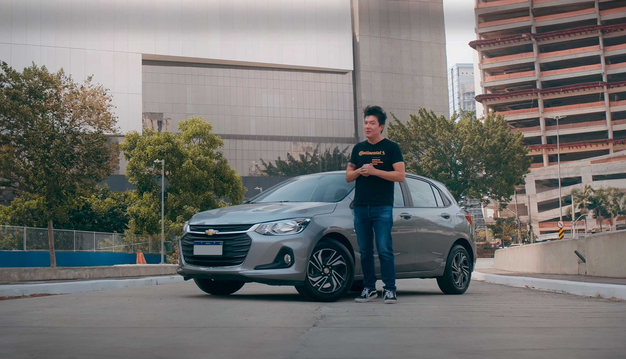 (Juliano Barata, do canal automotivo Flatout Brasil, um homem branco de cabelo curto preto em pé ao lado de um Chevrolet Onix 1.0 prata em um ambiente urbano. Ele veste uma camiseta preta com a logo da Continental. Ao fundo, há árvores e prédios altos, um deles em construção. A imagem sugere uma análise do veículo e seus pneus, com foco em manutenção e escolha dos pneus ideais para o Chevrolet Onix. O carro, destacado no cenário, está equipado com rodas esportivas e pneus UltraContact da Continental, e o homem parece pronto para discutir as vantagens de performance e segurança e desvantagens do modelo do veículo que é um dos mais vendidos do Brasil)