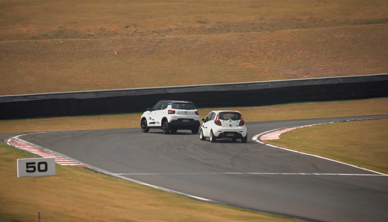 (Dois carros compactos brancos fazem uma curva fechada em uma pista de corrida. À esquerda, um Citroën C3 you com design robusto segue de perto um JAC J2 Turbo, um modelo hatch compacto. A pista apresenta marcações claras, com áreas de escape nas bordas. O número '50' está visível ao lado da pista, indicando a proximidade de uma curva acentuada. A imagem destaca o teste comparativo entre os veículos, onde os pneus Continental UltraContact são avaliados por sua performance em curvas fechadas e alta aderência, garantindo controle e segurança em situações desafiadoras.)