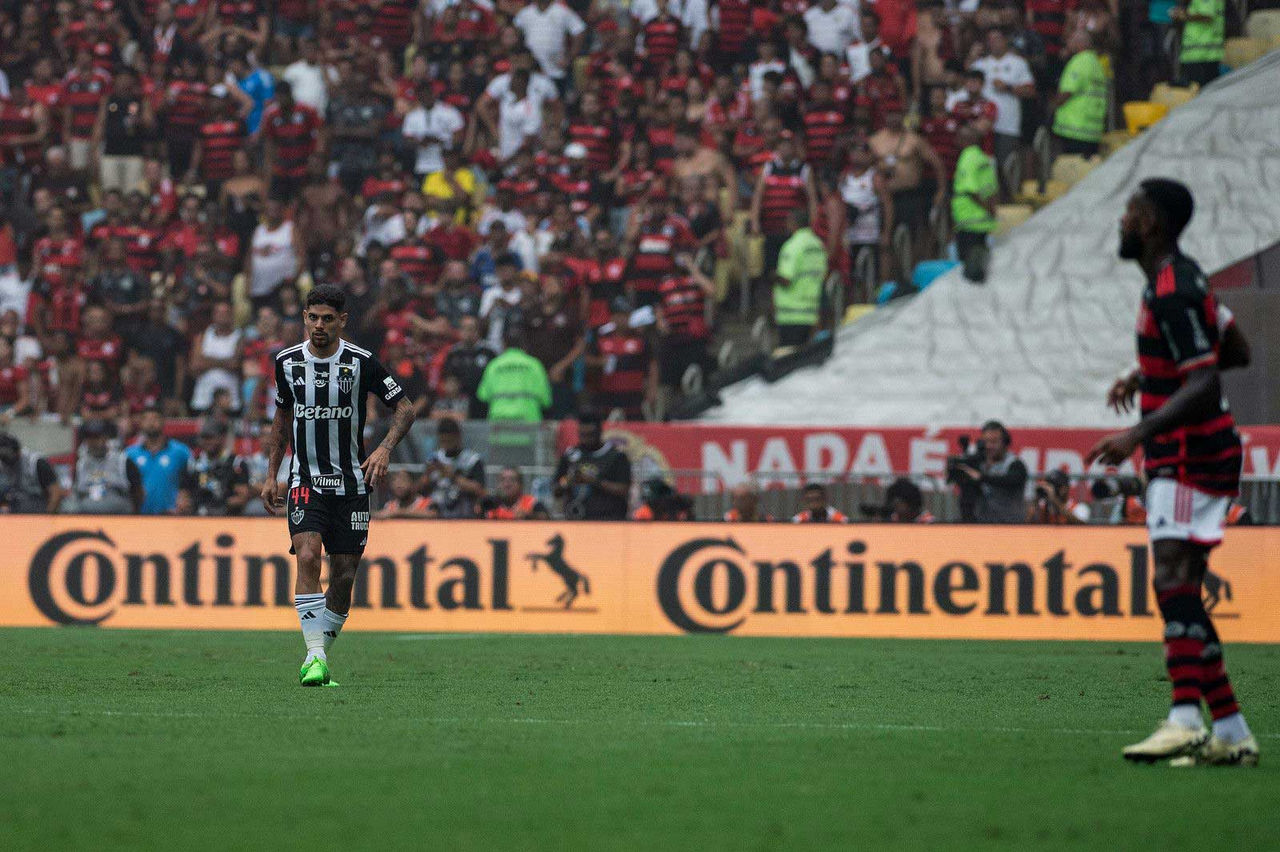 (A imagem retrata um momento de um jogo de futebol em um estádio cheio. Ao fundo, uma grande multidão de torcedores usa camisas vermelhas e pretas, típicas de um time de futebol, criando um ambiente vibrante e animado. Em primeiro plano, um jogador está caminhando em direção ao centro do campo. Ele veste um uniforme de cores claras e escuras com listras verticais, e seu número é 44. O campo é verde, e o clima parece abafado, evidenciado por algumas pessoas com roupas leves na arquibancada. Atras do jogador, há um painel de publicidade com a palavra "Continental". A atmosfera é de expectativa e tensão típica de uma partida importante)