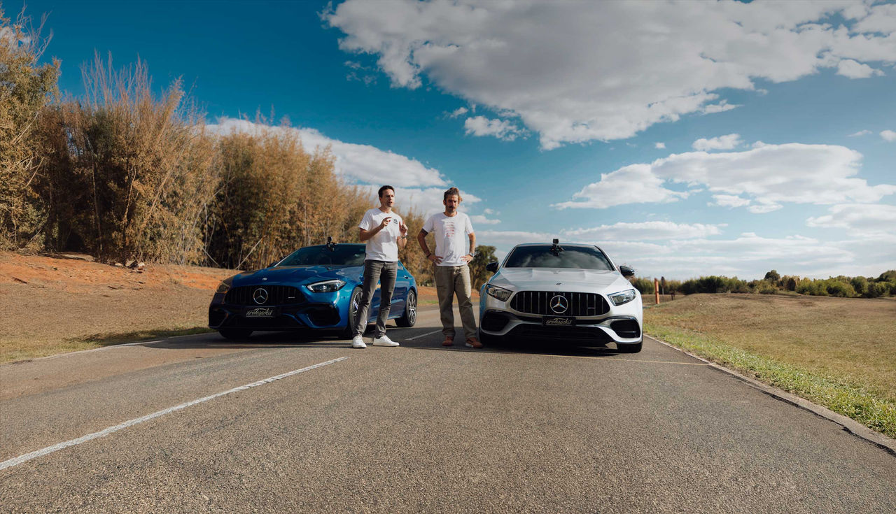 (A imagem retrata dois homens brancos de cabelos claros em pé ao lado de dois carros de luxo em uma estrada. À esquerda, um Mercedes AMG C 63 S E Performance azul, enquanto à direita, um AMG 45 S 4Matic branco. Os homens estão vestidos de maneira casual, usando camisetas claras, ambos com calças. O ambiente ao redor é um espaço aberto, com uma vegetação baixa e algumas árvores ao fundo. O céu está parcialmente nublado, com nuvens brancas flutuando. A atmosfera é descontraída, sugerindo um momento de apreciação dos carros e do cenário)
