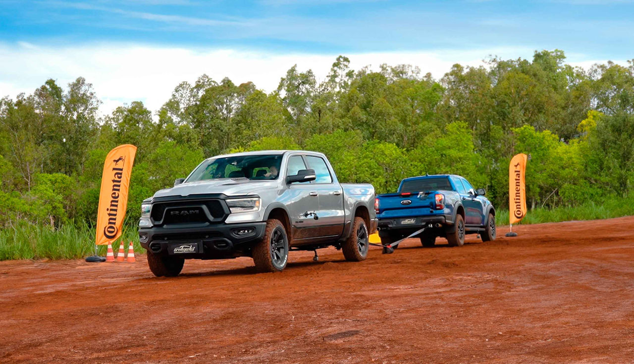 (A imagem mostra dois grandes veículos pickup parados lado a lado em um terreno de terra vermelha. O carro à esquerda é de cor prata e tem a marca RAM claramente visível na grade dianteira. À direita, há um pickup de cor azul que apresenta um design robusto e esportivo. Atrás dos veículos, o ambiente é verdejante, com árvores e vegetação que sugerem uma área rural ou um campo aberto. Ao fundo, o céu é azul com algumas nuvens dispersas. No primeiro plano, do lado esquerdo e direito da imagem, há duas bandeiras verticais na cor laranja com o logotipo da Continental e a silhueta de um cavalo, indicando possivelmente um evento ou uma promoção da marca de pneus. Não há pessoas visíveis na imagem, colocando todo o foco nos veículos e no cenário ao redor deles.)