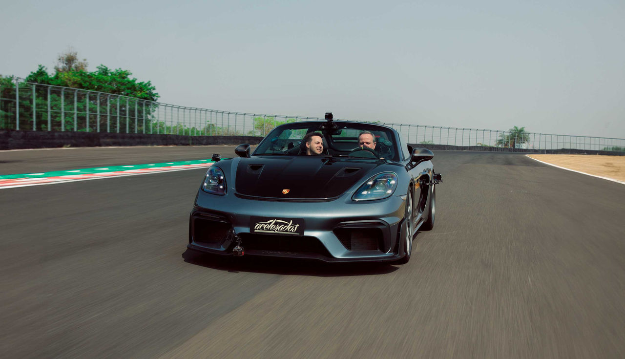 (Você está vendo o carro esportivo de cor cinza escuro, modelo Porsche 718 Spyder RS, com o capô dianteiro voltado para a câmera. O veículo está em movimento em um circuito de corrida, onde vemos marcas de zebra (branco com listras vermelhas e verdes) ao longo da borda da curva da pista. Duas pessoas estão no carro, Gerson e Rubinho, um passageiro e um motorista, ambos usando óculos de sol e com expressões animadas. A cabeça do passageiro está inclinada para a frente, indicando velocidade e movimento. Ao fundo, você pode ver uma cerca alta de metal com vegetação logo atrás dela, além de uma árvore isolada e o céu claro. É uma cena que transmite ação e emocionante performance de condução. O teste utiliza pneus Continental SportContact 7, conhecidos pela aderência e durabilidade. O modelo conversível e aerodinâmico do Porsche 718 Spyder RS é projetado para máxima performance em pistas)