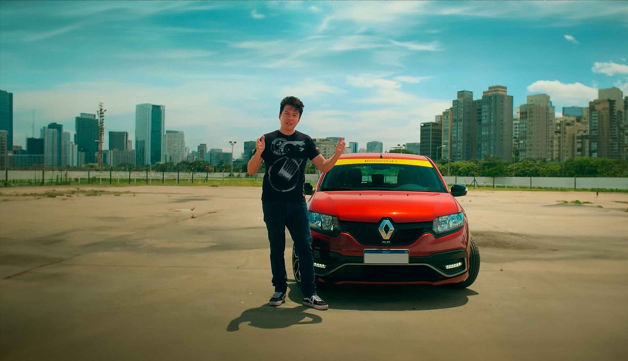 (Apresentador Juliano Barata gesticula em frente a um Renault Sandero R.S. vermelho equipado com pneus Continental, com faixa amarela no para-brisa destacando a marca Continental Pneus. A imagem foi capturada em um ambiente urbano ao ar livre, com prédios altos ao fundo, durante gravação sobre calibragem correta dos pneus e seus impactos na segurança e desempenho