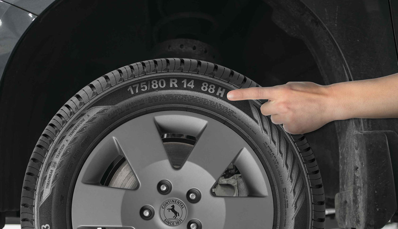 (uma mão branca apontando para a lateral de um pneu Continental instalado em um veículo, destacando o índice de velocidade 'H' da medida “175/80 R 14 88H” gravado na borracha. Ao centro da roda é possível ver o logotipo da marca Continental. O índice de velocidade do pneu indica a capacidade máxima de velocidade que ele pode suportar com segurança, sendo essencial para escolher o pneu correto para o seu veículo. O código 'H' representa uma velocidade máxima de 210 km/h. O fundo da imagem é um ambiente típico de oficina, com algumas ferramentas e equipamentos ao redor, mas não muito detalhados. A iluminação parece ser adequada, permitindo que os elementos do pneu e da roda se destaquem claramente)