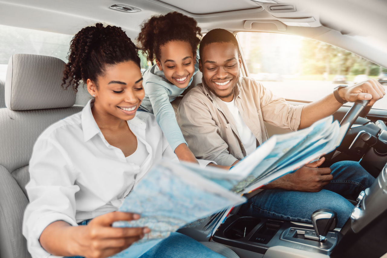 Cheerful African American Family Looking At Road Map Sitting In Car. Parents And Daughter Choosing Destination For Summer Road Trip Together. Local Tourism Concept. Selective Focus