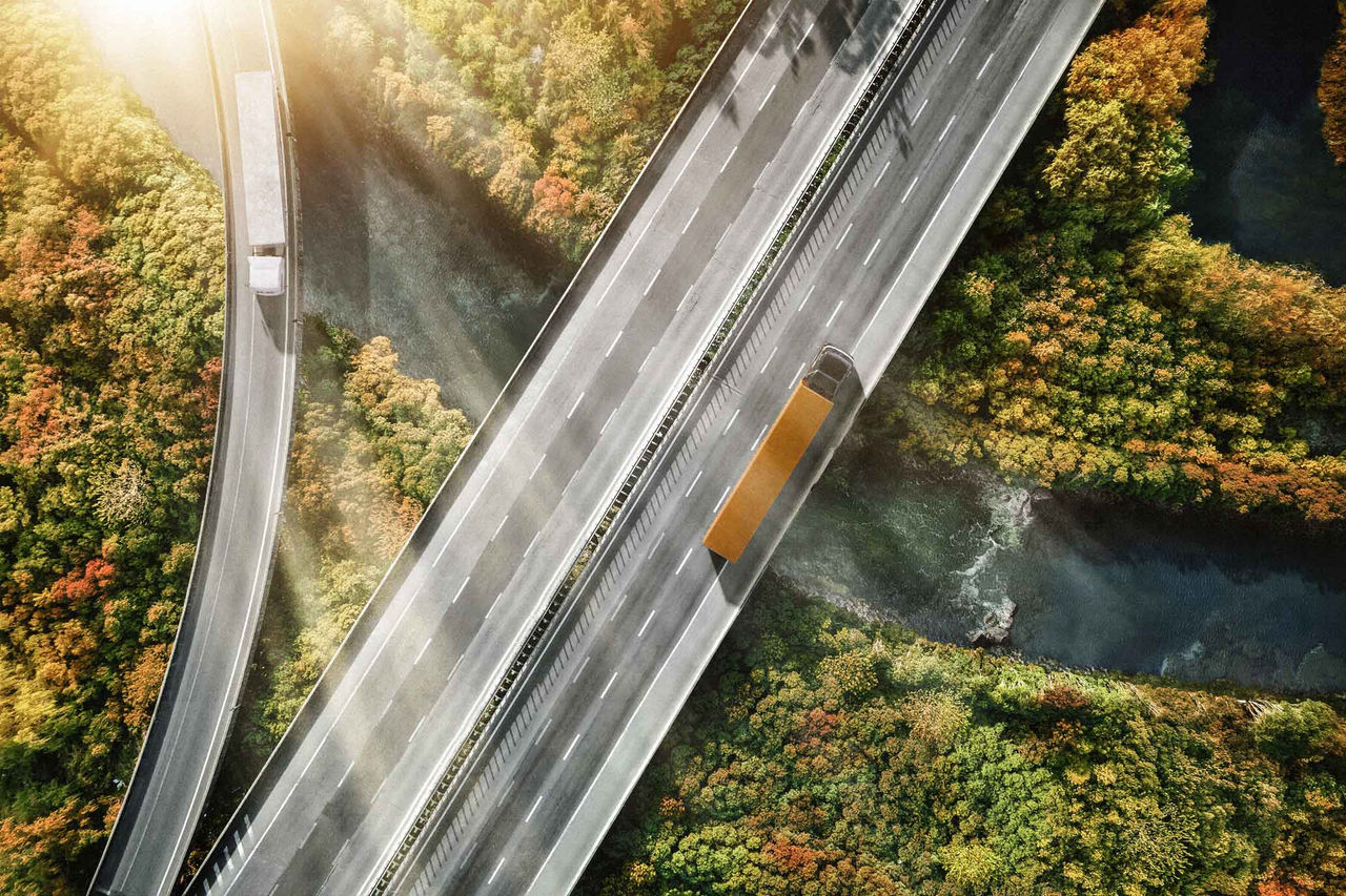 (Mostra uma vista aérea de uma rodovia que corre sobre um rio sinuoso e cercada por vegetação. Há duas faixas de tráfego visíveis; uma delas, à direita, está ocupada por um caminhão longo de cor predominantemente cinza com uma caçamba laranja. A rodovia apresenta um asfalto claro, e as faixas são demarcadas com linhas brancas. Ao redor, a paisagem é rica em tonalidades de verde, laranja e amarelo, sugerindo que é outono. O rio abaixo é de um tom mais escuro e serpenteia suavemente entre a vegetação, enquanto a luz do sol cria um brilho nas partes superiores da imagem, indicando que é um dia claro)