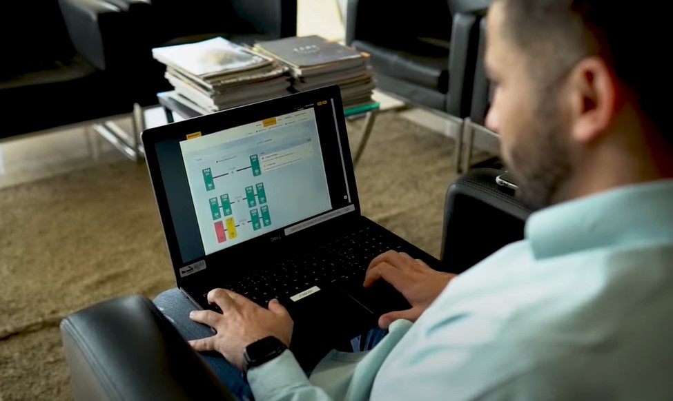 (foto de homem branco com barba e camisa verde, sentado em sofá com notebook no colo - aplicativo do sistema ContiConnect aparece na tela - ao fundo poltronas e mesa com revista)
