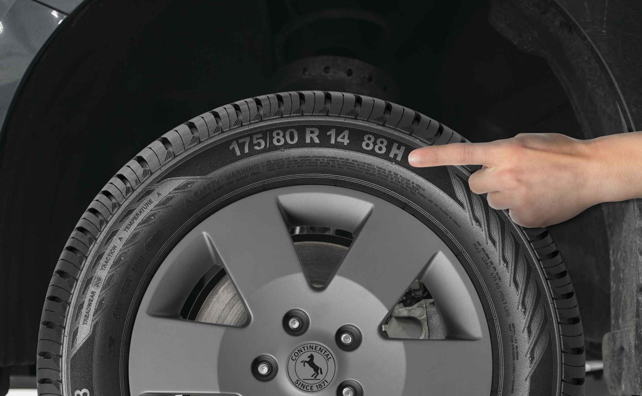 (uma mão branca apontando para a lateral de um pneu Continental instalado em um veículo, destacando o índice de velocidade 'H' da medida “175/80 R 14 88H” gravado na borracha. Ao centro da roda é possível ver o logotipo da marca Continental. O índice de velocidade do pneu indica a capacidade máxima de velocidade que ele pode suportar com segurança, sendo essencial para escolher o pneu correto para o seu veículo. O código 'H' representa uma velocidade máxima de 210 km/h. O fundo da imagem é um ambiente típico de oficina, com algumas ferramentas e equipamentos ao redor, mas não muito detalhados. A iluminação parece ser adequada, permitindo que os elementos do pneu e da roda se destaquem claramente)