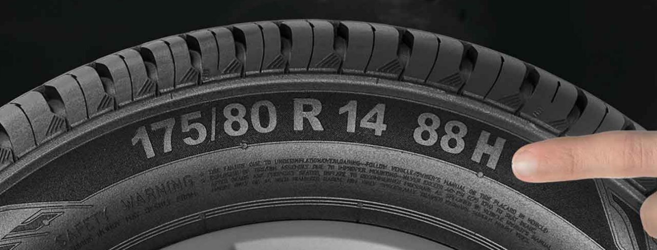 (uma mão branca apontando para a lateral de um pneu Continental instalado em um veículo, destacando o índice de velocidade 'H' da medida “175/80 R 14 88H” gravado na borracha. Ao centro da roda é possível ver o logotipo da marca Continental. O índice de velocidade do pneu indica a capacidade máxima de velocidade que ele pode suportar com segurança, sendo essencial para escolher o pneu correto para o seu veículo. O código 'H' representa uma velocidade máxima de 210 km/h. O fundo da imagem é um ambiente típico de oficina, com algumas ferramentas e equipamentos ao redor, mas não muito detalhados. A iluminação parece ser adequada, permitindo que os elementos do pneu e da roda se destaquem claramente)