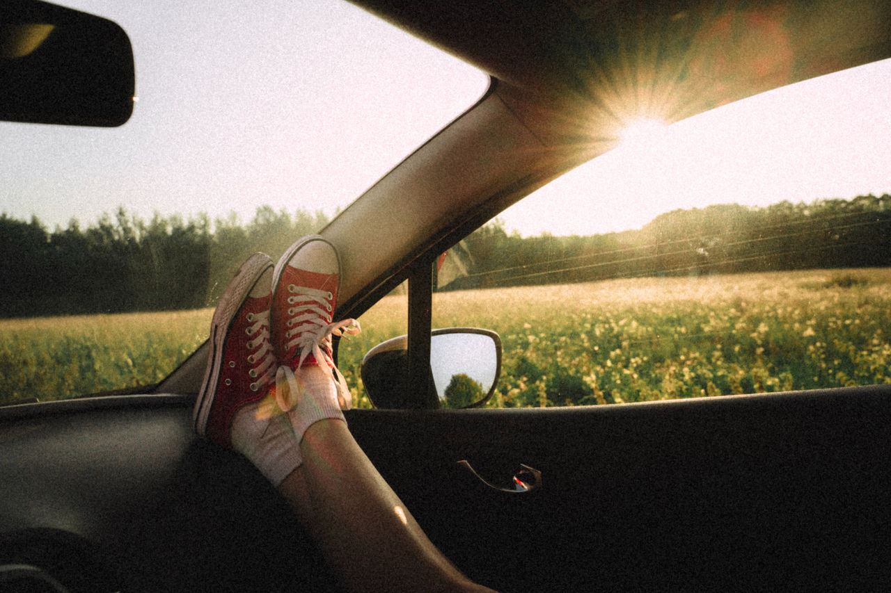 (foto interna do carro com pessoa com pé apoiados em painel do carro e por do sol aparecendo na janela)